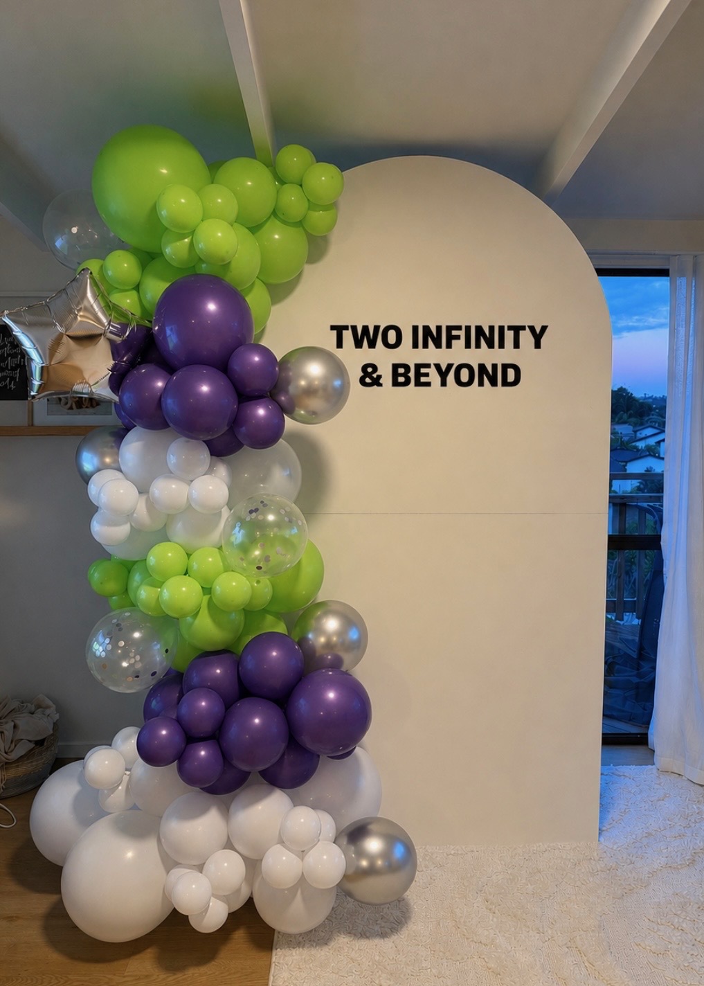 Second birthday setup — cream arch backdrop with custom vinyl lettering and a lime green, purple, white and silver balloon garland with confetti balloons and a silver star foil
