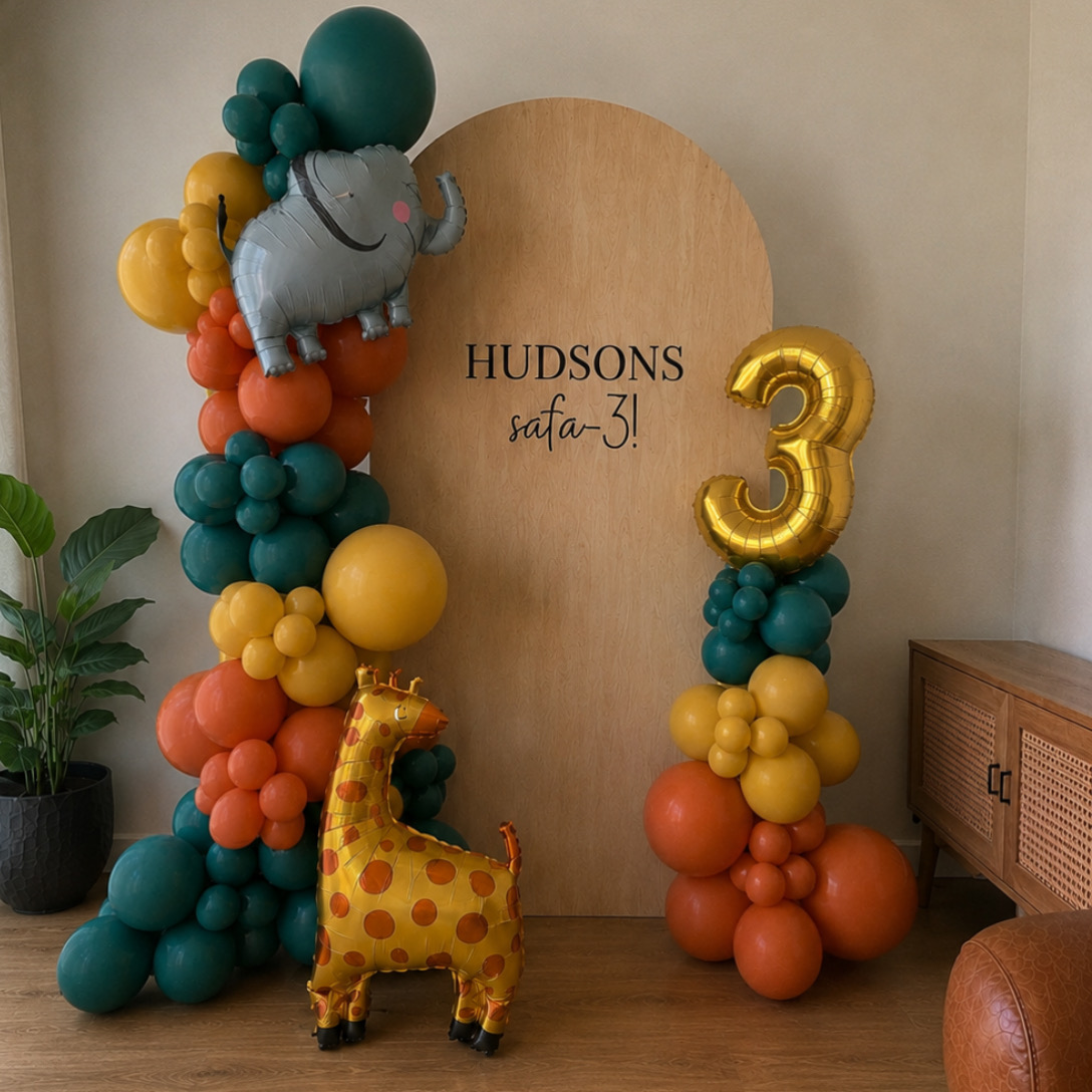 Safari-themed third birthday setup — plywood arch backdrop with custom lettering, teal, mustard and orange balloon garland with elephant and giraffe foils, and a freestanding column topped with a gold number three